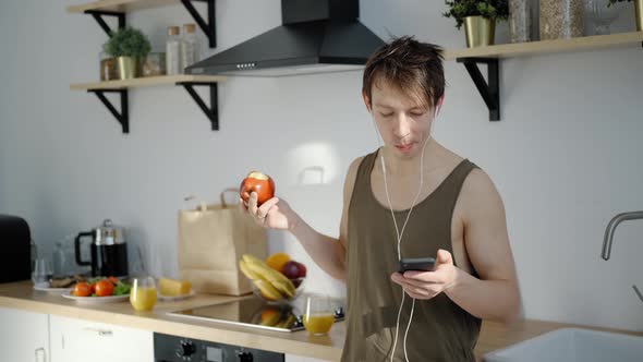 Cheerful Young Man Eating Apple, Listening Music in Light Kitchen