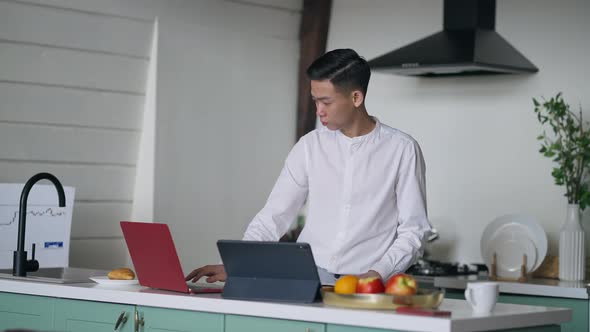 Concentrated Serious Asian Man Analyzing Document Typing on Laptop Keyboard and Scrolling Tablet alt