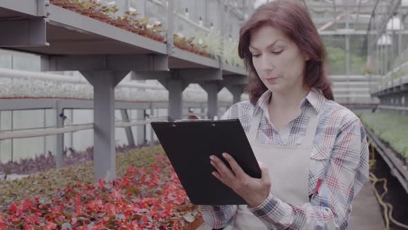 Portrait of Busy Caucasian Woman Taking Notes and Smiling at Camera in Greenhouse. Mid-adult alt