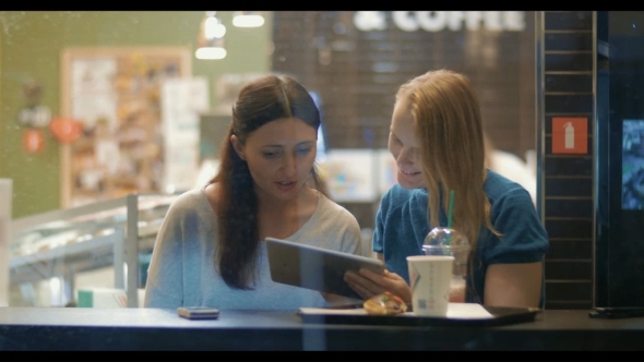 Female Friends Talking And Using Touch Pad In Cafe Female Friends Talking And Using Touch Pad In Cafe