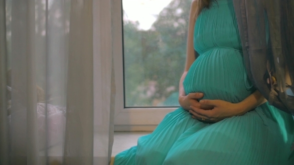 Pregnant Woman Sitting On Windowsill And Embracing Pregnant Woman Sitting On Windowsill And Embracing