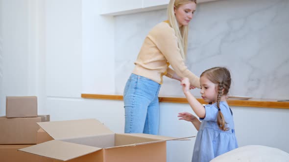 Funny little girl with long plaits unpacks brown cardboard box and gives mother white plates alt