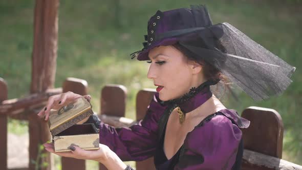 Side View Beautiful Lady in Halloween Witch Costume Opening Gift Box with Surprised Facial alt