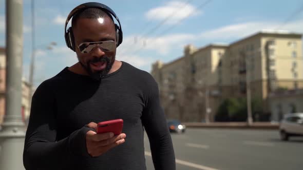 Portrait of an African Man in Dark Clothing, Sunglasses and Headphones. He Holds a Red Phone in His alt