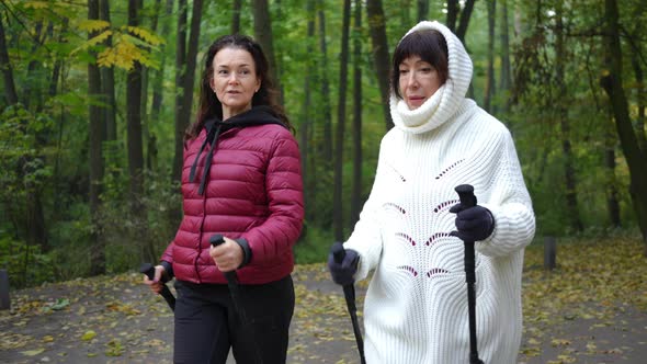 Two Mature Caucasian Women Talking Walking with Poles in Autumn Park Working Out alt