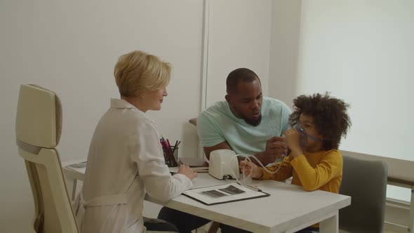 Cute Ill Preschool African American Boy Making Inhalation with Nebulizer at Medical Clinic alt