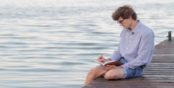 Man Writes In A Notebook Sitting By The River