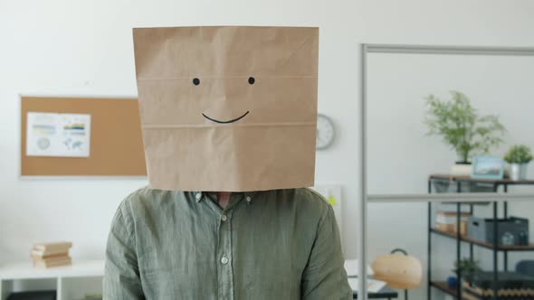Portrait of Male Employee Wearing Paper Bag with Smile Emoji Standing in Office and Looking at alt