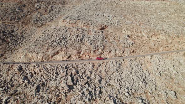 Aerial drone side shot of red car driving along the narrow coastal road above rocky shore alt