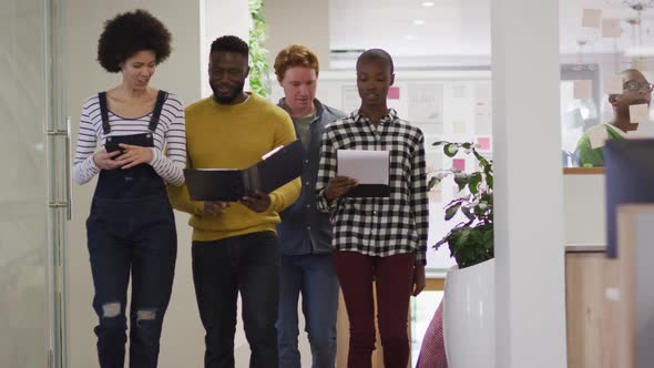 Diverse smiling male and female business colleagues walking and discussing paperwork in office alt