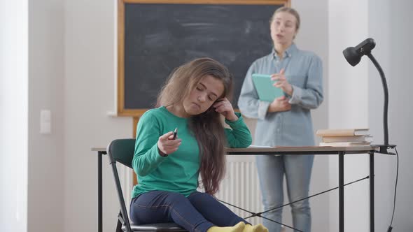 Bored Little Person Sitting on Chair at Desk in Classroom Playing with Pen and Crossing Hands As alt