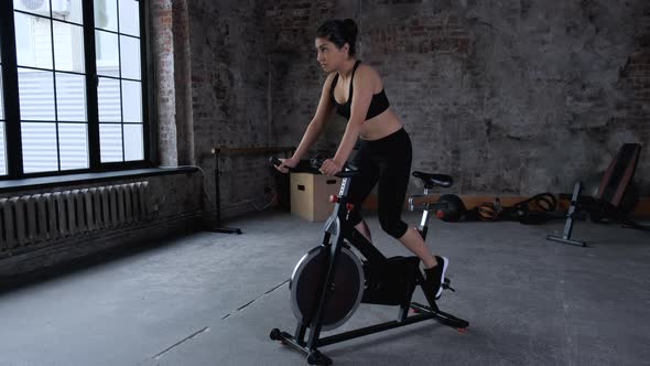 Focused young Indian woman cycling in gym. Side view of slim brunette sportswoman alt