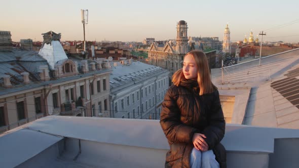 Teen Girl In Jacket Is Sitting On The Roof Of Old European City At Sunset