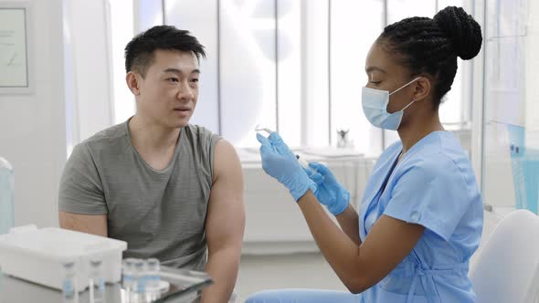Young Man Communicating with Doctor in Protective Mask and Safety Gloves While She Filling Syringe alt