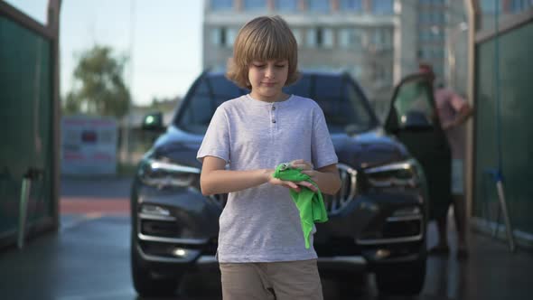 Boy Cleaning Hands Standing at Blurred Black Vehicle with Man Walking at Background alt