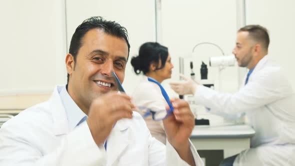 Doctor Puts on Glasses and Shows Thumbs Up While on the Background Another Doctor Examines the