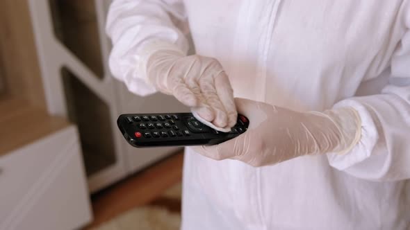 Woman in a Protective Suit Wiping the Remote Control with a Disinfecting Cloth. alt