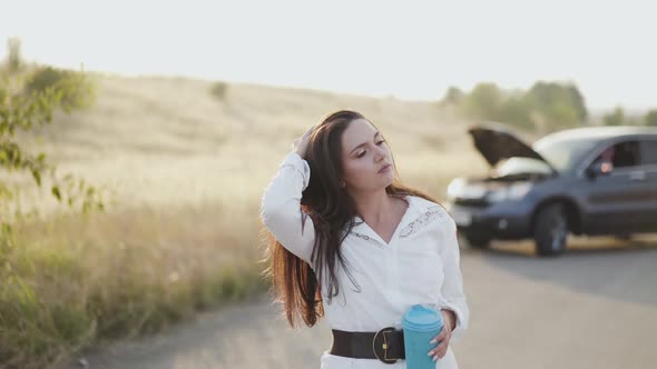 Girl with Cup in Hand Corrects Hair Talks and Looks Around at Broken Car alt