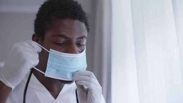Male Doctor in Surgical Gloves Putting on Coronavirus Face Mask Standing in Hospital Indoors alt