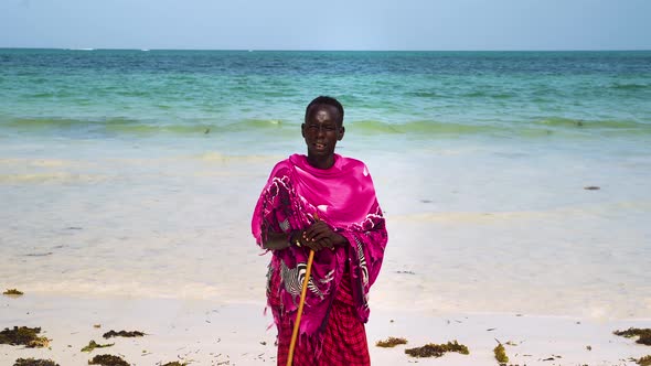 African maasai man in pink clothing standing on beach leaning on stick. alt