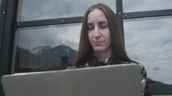 A Young Freelancer Girl Works at a Laptop in a Cafe Near the Mountains alt