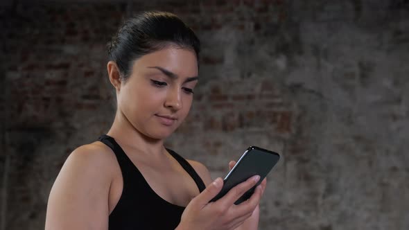 Indian young woman using smart phone to surf internet in gym after workout. smiling female chats. alt