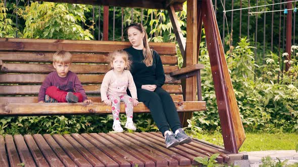 Travelling: Young Pretty Mother with Children, Swinging on a Wooden Swing Near the Summer House alt