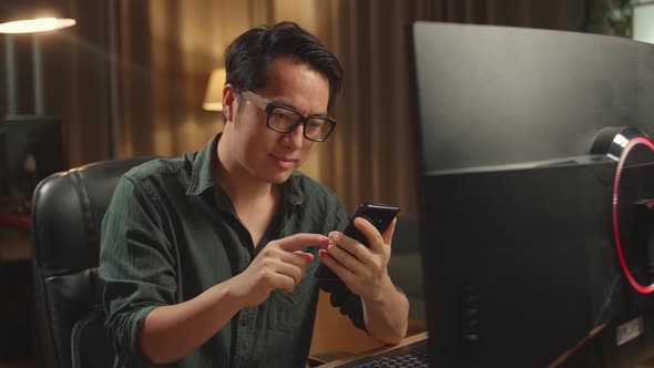 Asian Male Using Mobile Phone While Works On His Personal Computer With Big Display alt
