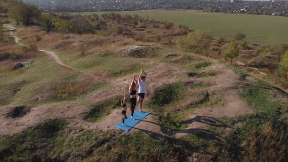 Orbiting Aerial Shot of Family of Three Mother Father and Daughter Do Yoga Exercises on Top of Hill alt