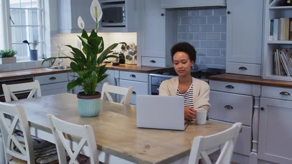 Mixed race woman using laptop and drinking coffee in kitchen alt