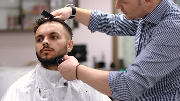 Unparalleled Barber with a Beard and a Tattoo Is Cutting the Hair of His Client in the Barbershop. alt