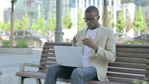 African Man Talking on Video Call While Sitting Outdoor on Bench alt
