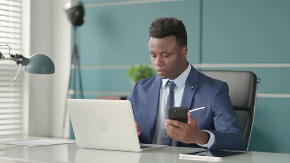 African Businessman Using Smartphone While Using Laptop in Office alt