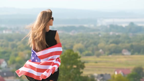 Young Pretty American Woman with Long Hair Holding Waving on Wind USA Flag on Her Sholders Standing alt