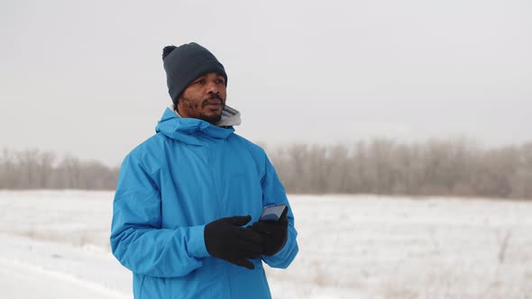 Black Man Stands Outdoor in Winter Clothes and Looks Into Smartphone Using GPS Application Front alt