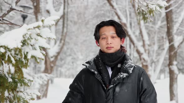 Young Asian Man in Winter Jacket Stands in Snowy Forest and Fixes His Hair alt