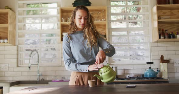 Happy caucasian woman standing at counter in cottage kitchen pouring tea from teapot and smiling alt