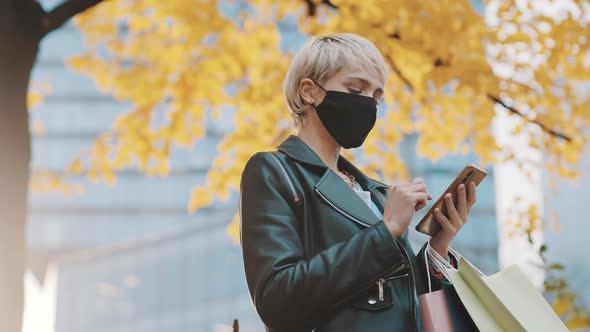 Young Woman with Face Mask and Shopping Bags Using Smartphone Under the Tree with Yellow Leaves alt