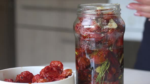 A Woman Pours Spices And Sun Dried Tomatoes Into A Jar With Olive Oil. alt