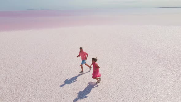 Aerial Drone Footage of Happy Young Couple in Pink Wear Having Fun and Joyfully Running on White alt