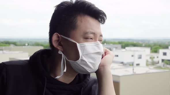 A Young Asian Man in a Face Mask Stands on a Balcony and Looks at a Cityscape Below alt
