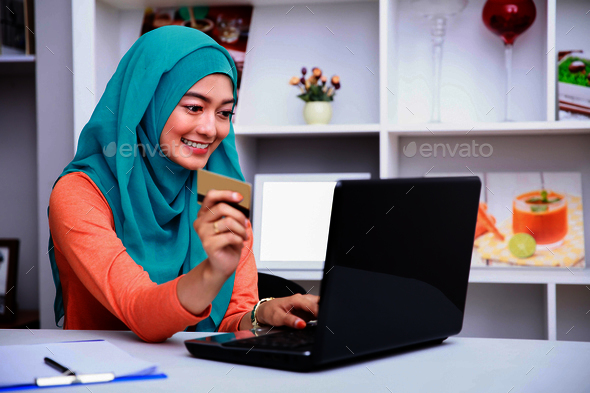 young muslim woman browsing internet on laptop while holding a c