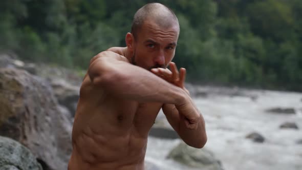 an Unshaven Man with a Bare Torso is Boxing By a Mountain River alt