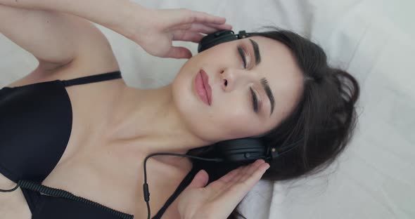 Woman on Bed Listens to Music on Headphones Opens Eyes and Smiles Into Camera alt