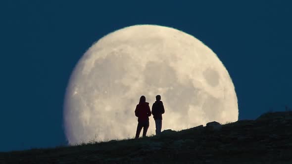 Silhouette of Two People Standing Against Huge Moon Background.