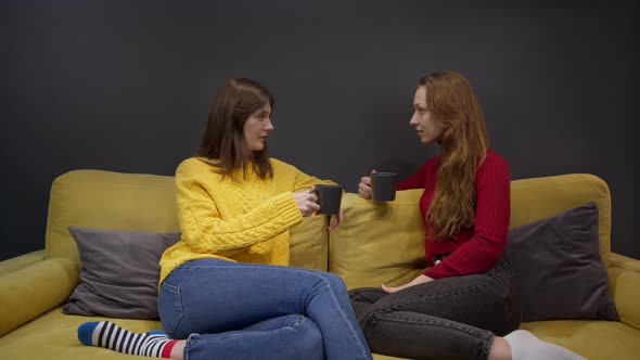 Two Girls Discussing Gossip Sitting on Yellow Couch and Drinking Coffee