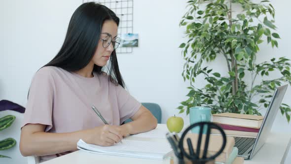 Young Asian Lady Looking at Laptop Screen and Writing Doing Homework Indoors in Apartment alt