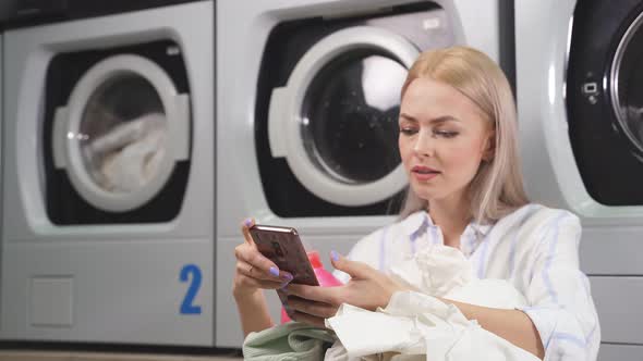 Portrait of a Beautiful Young Blonde Sitting in the Laundry Room Next to the Washing Machine and