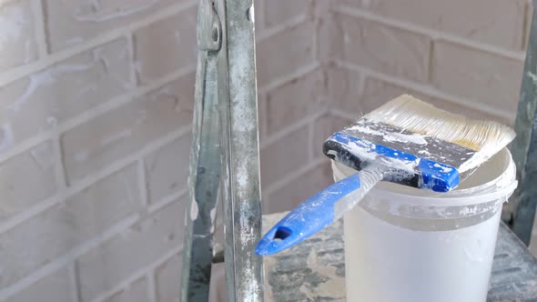 A hand in a work glove dips a paint brush in white paint in a bucket on a ladder against a brick wal alt