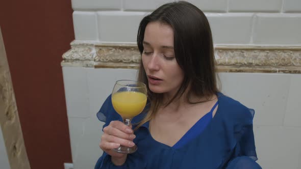 A Beautiful Girl in a Room in a Blue Dress Sniffs and Then Enjoys Drinking Orange Juice From a Wine